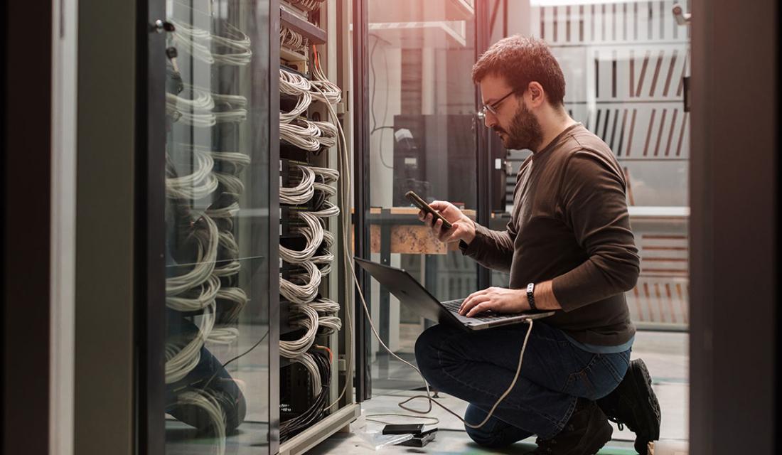 Ein Mann kniet ein einem Serverraum vor einem Schrank und liest Daten aus